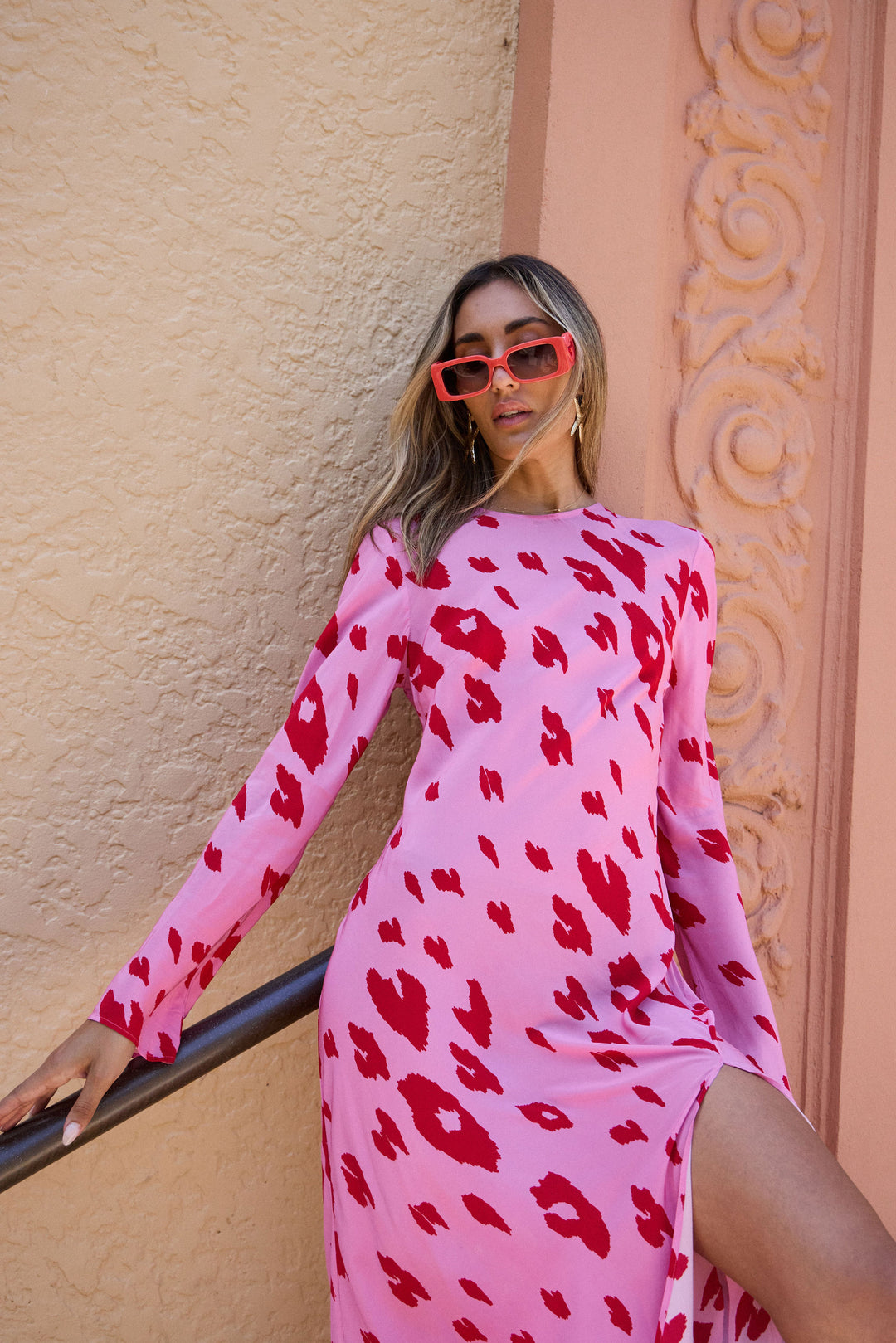 Woman wearing a pink dress with red patterns and red sunglasses against a beige wall.