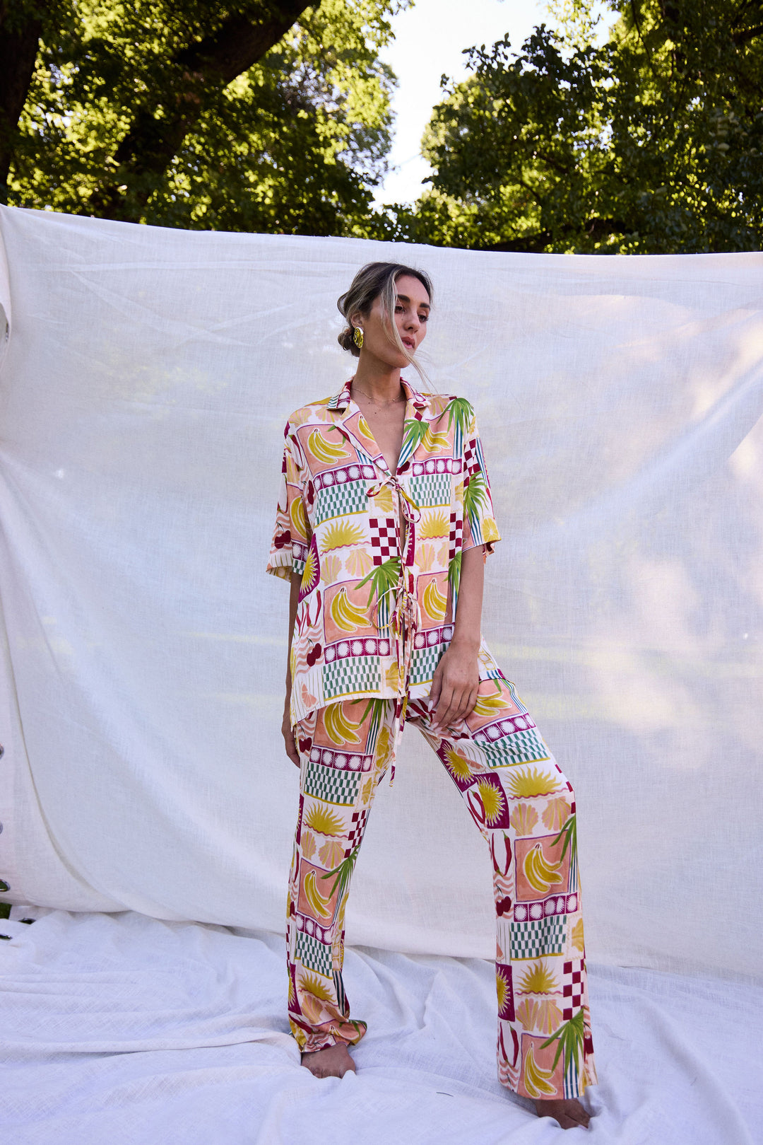 Person wearing a colorful patterned outfit standing against a white backdrop with trees in the background.