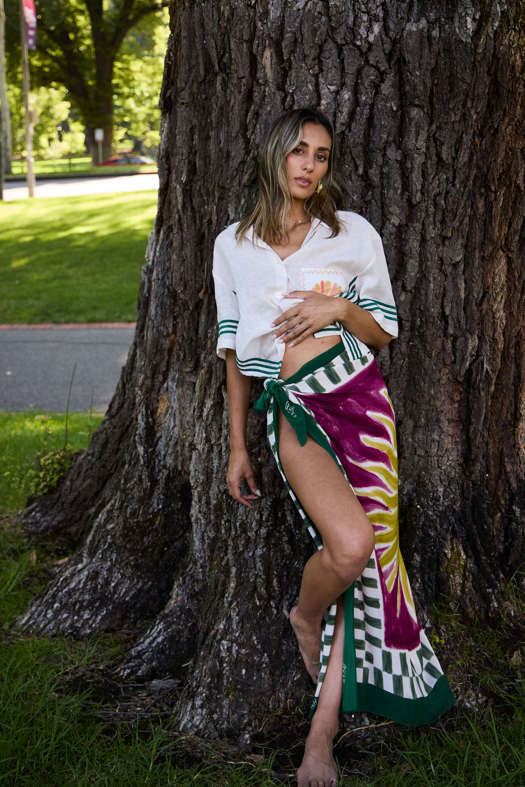 Woman in a white shirt and colorful skirt standing next to a large tree in a park.