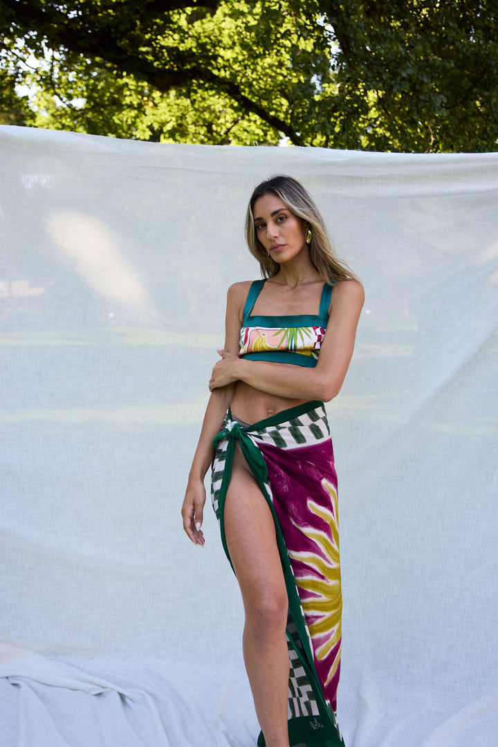 Woman wearing a colorful bikini and patterned sarong against a white backdrop with greenery.