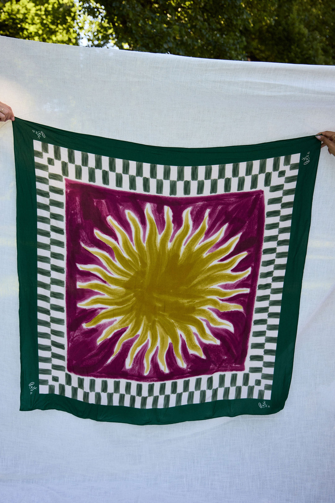 Scarf with a sun design on a green border held by two hands against a natural background.