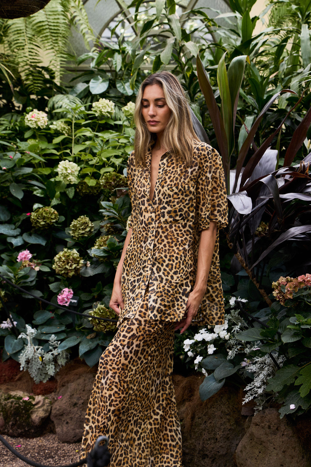 Woman wearing a leopard print outfit standing among lush greenery and flowers.