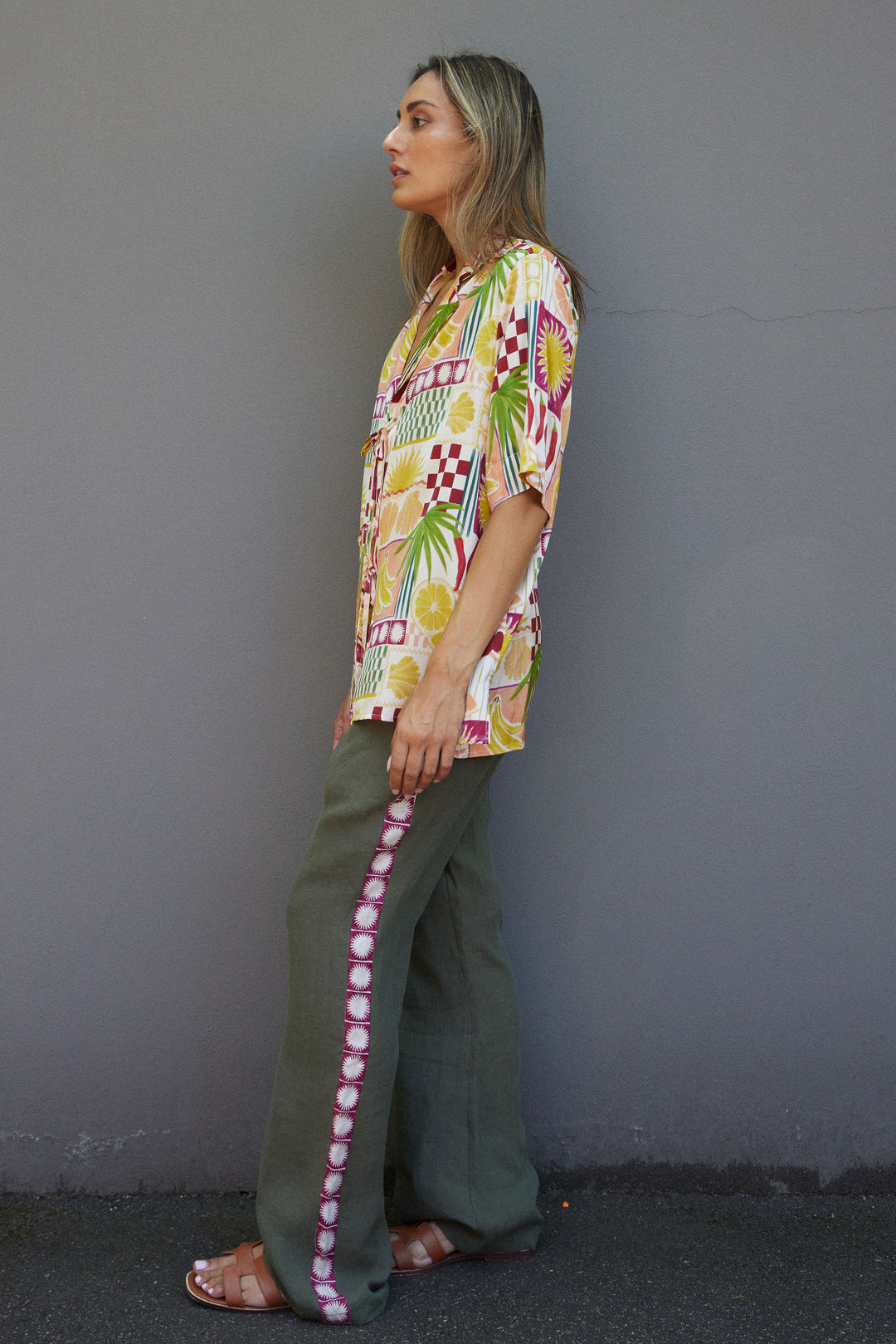 Woman wearing a colorful patterned shirt and green pants against a gray wall.