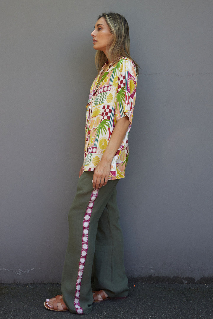 Woman wearing a colorful patterned shirt and green pants against a gray wall.