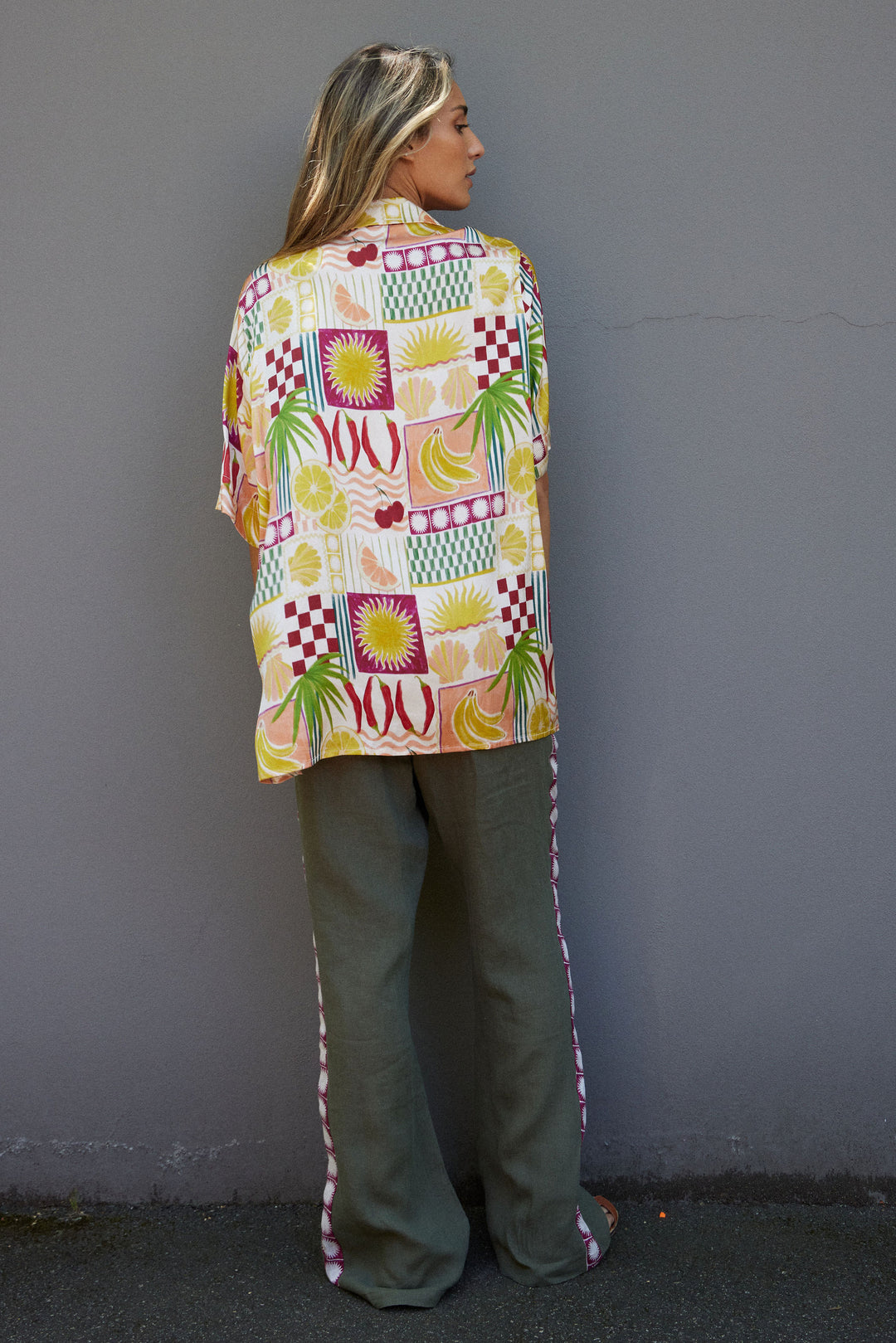 Person wearing a colorful patterned shirt and green pants against a gray wall.