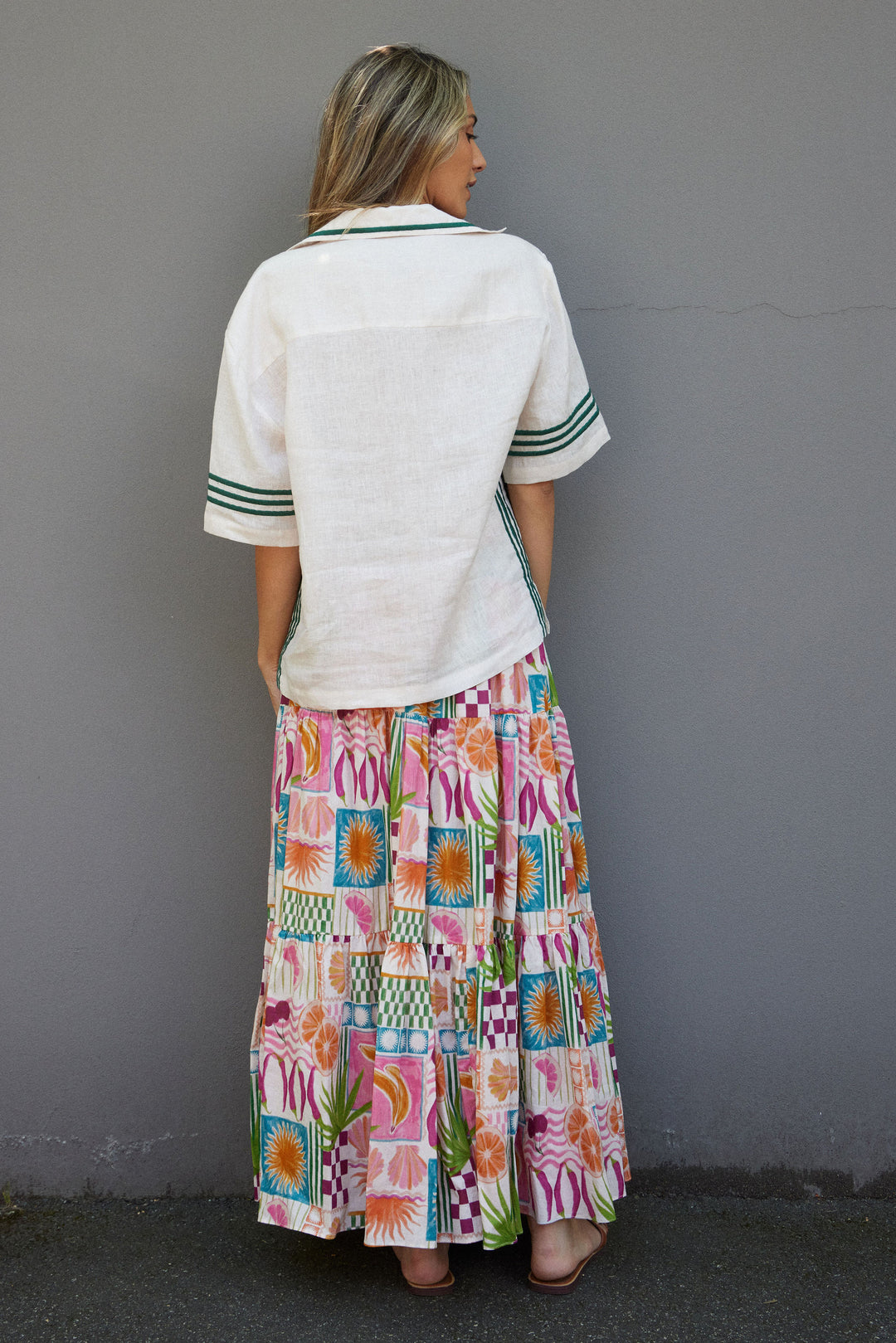 Woman wearing a white top with green trim and a colorful patterned skirt against a gray wall.