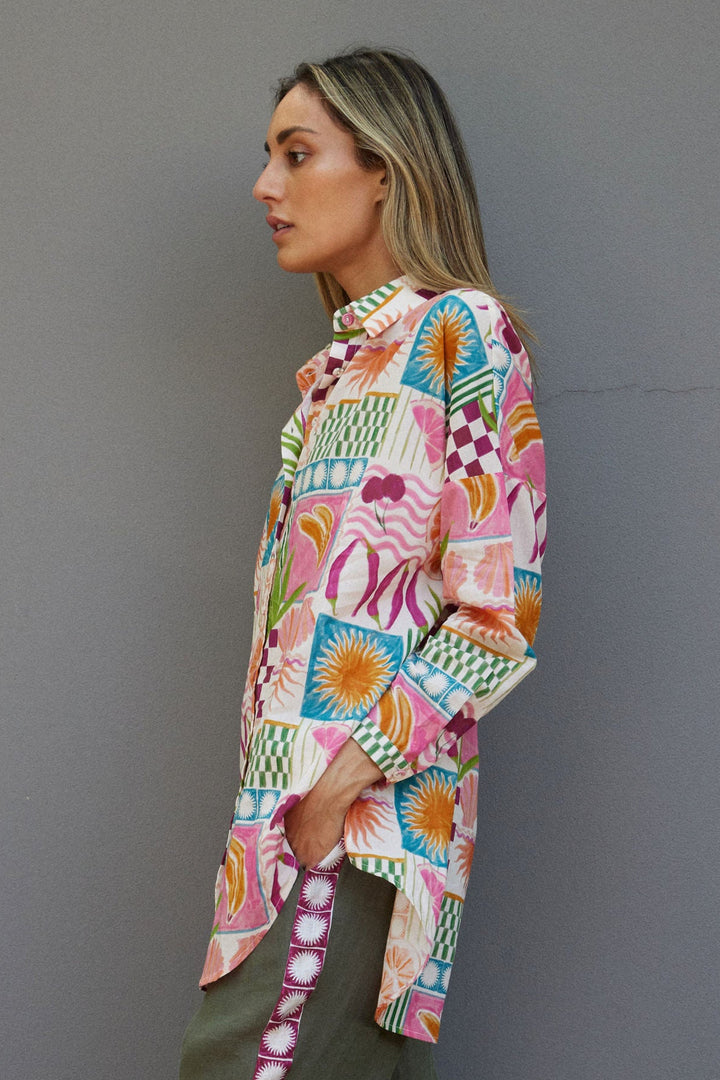 Woman wearing a colorful patterned shirt and green pants against a gray wall.