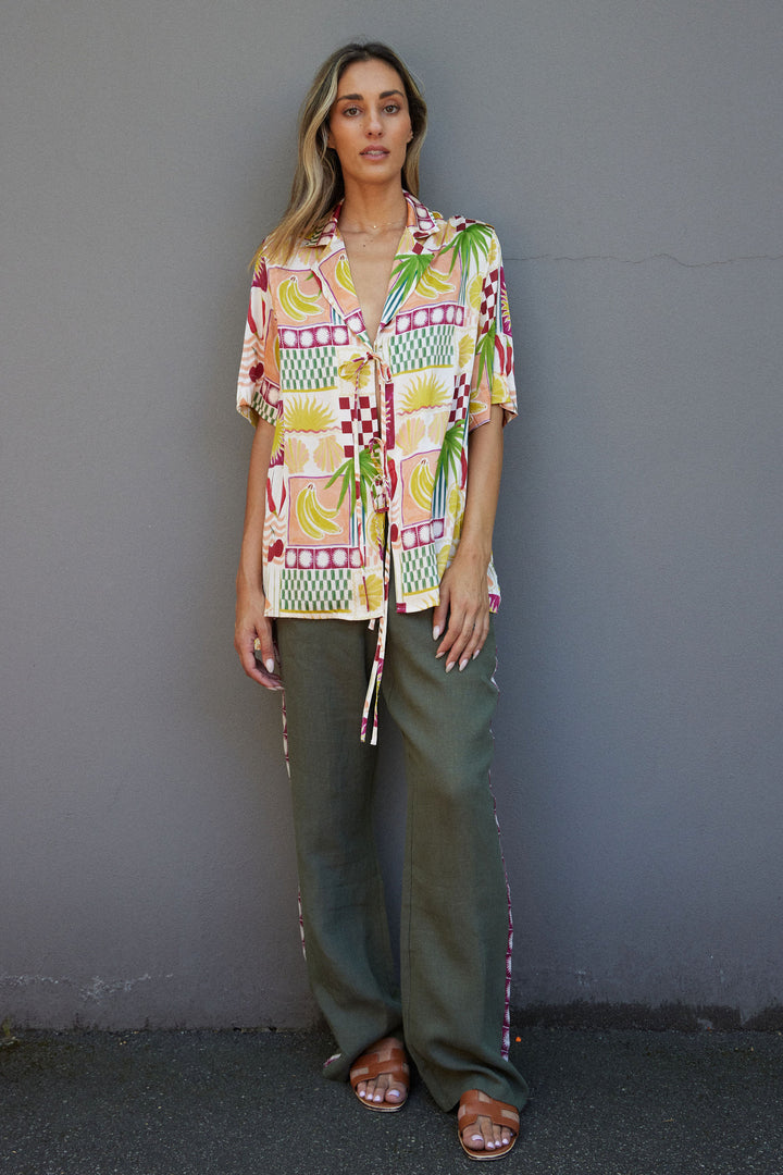 Woman wearing a colorful patterned shirt and green pants against a gray wall.